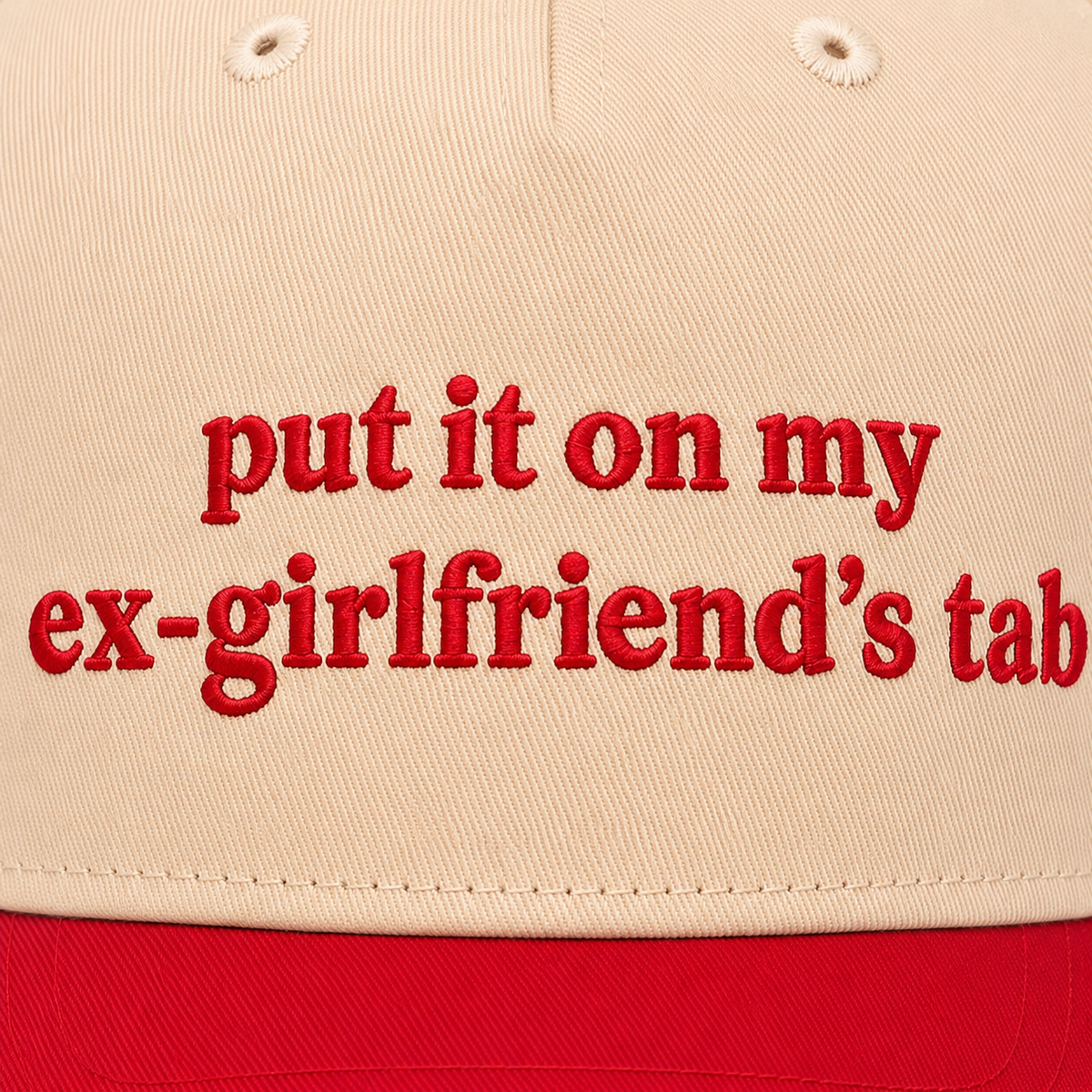 Close-up of bold red embroidered "put it on my ex-girlfriend's tab" lettering on a cream baseball cap front panel with red brim.
