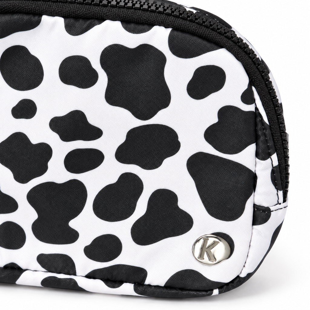 Close-up of Cow Print Belt Bag showing white nylon with large black cow print spots, silver Katydid K logo rivet, and black zipper edge detail
