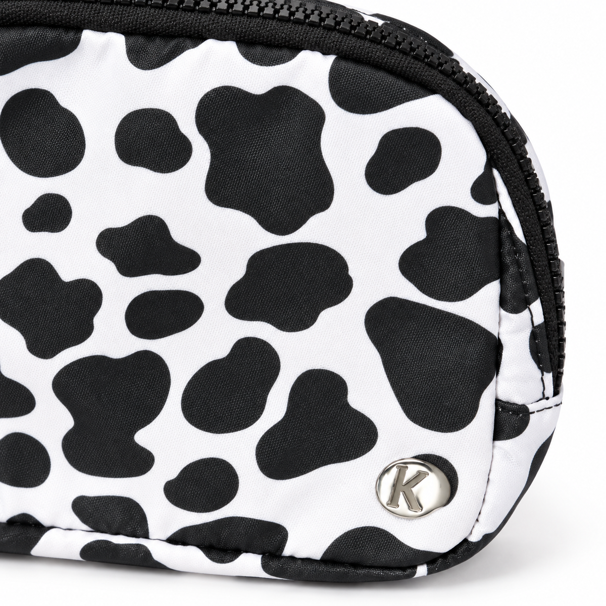 Close-up of Cow Print Belt Bag showing white nylon with large black cow print spots, silver Katydid K logo rivet, and black zipper edge detail
