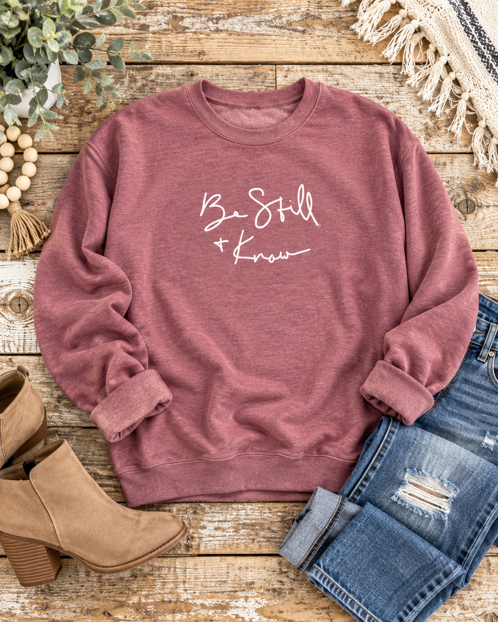 Dusty mauve Be Still + Know sweatshirt styled with distressed jeans and tan ankle boots on a rustic wood background

