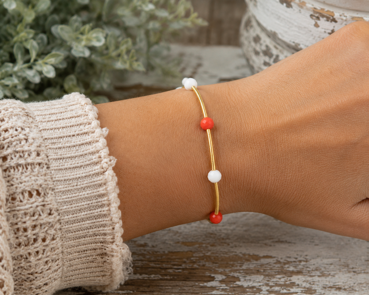 Woman wearing gold coil stretch bracelet with red and white bead accents on wrist with cream crochet knit sleeve and rustic white pedestal backdrop
