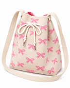 Coquette Bow Crossbody Bucket Bag three-quarter angle view showing cream woven knit fabric with allover pink bow pattern, tapered bucket silhouette wider at top, cream rope drawstring closure with silver aglet tips, and wide flat cream crossbody strap on white background
