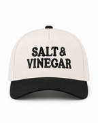 Salt & Vinegar Two-Tone Baseball Cap in Black front view showing cream structured cotton crown with bold black embroidered Salt & Vinegar block lettering, solid black bill, and black top button on white background
