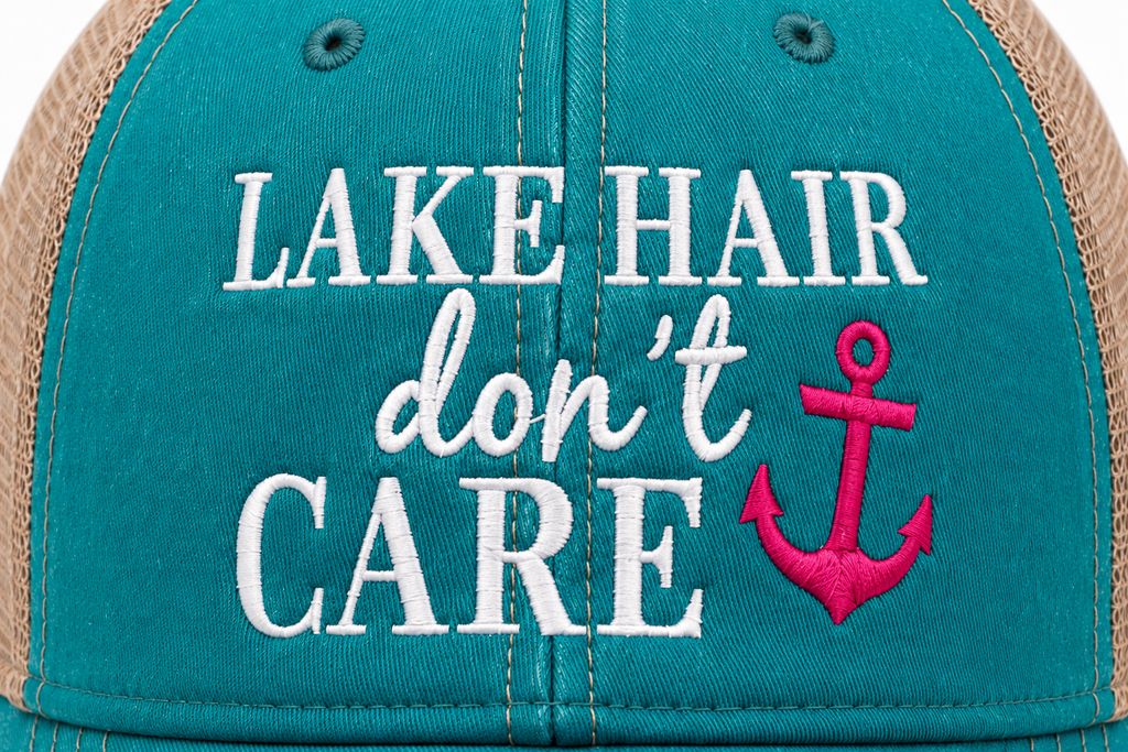 Close-up of a trucker hat front panel with teal fabric, tan mesh sides, and white embroidered text reading 'LAKE HAIR don't CARE' alongside a bright pink anchor graphic.