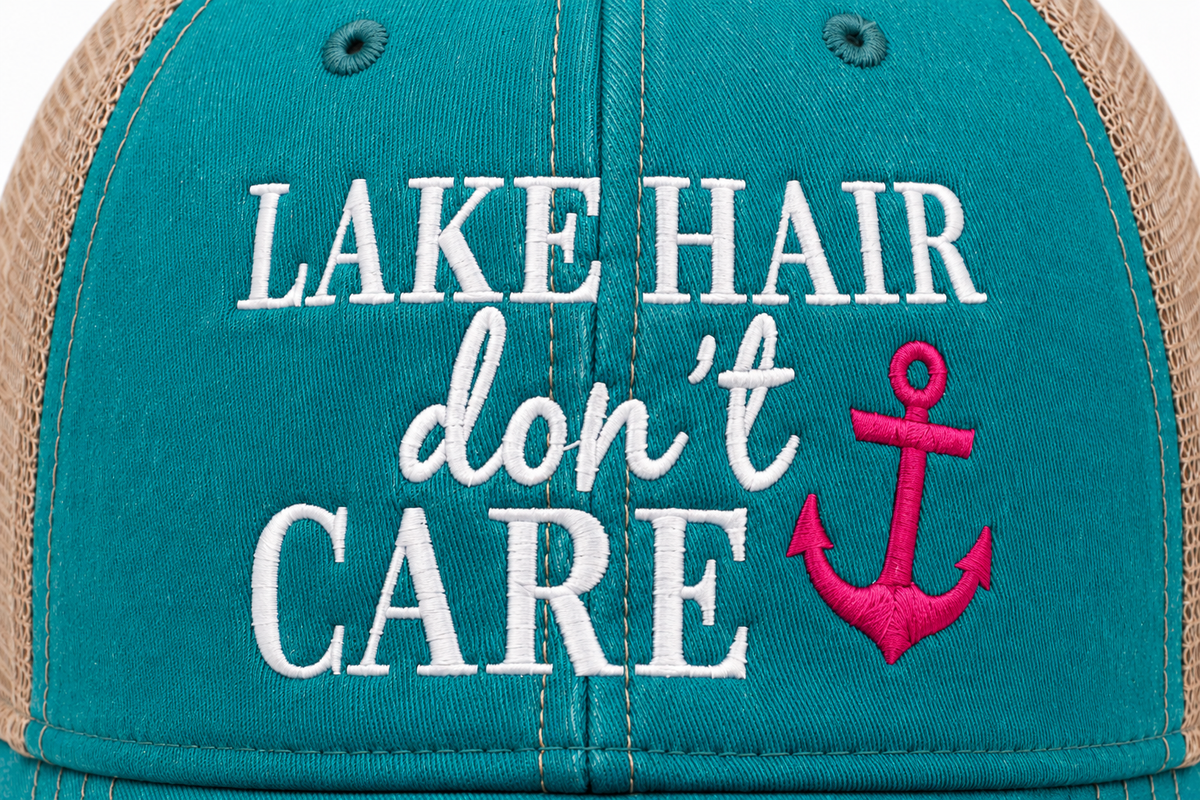 Close-up of a trucker hat front panel with teal fabric, tan mesh sides, and white embroidered text reading 'LAKE HAIR don't CARE' alongside a bright pink anchor graphic.