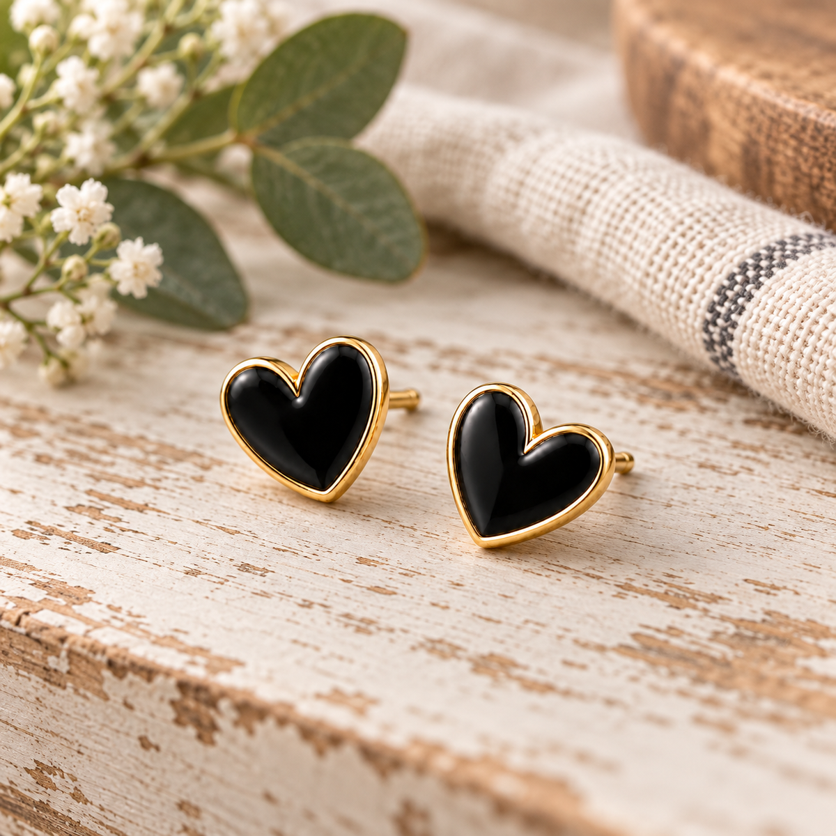 Black heart stud earrings with gold-tone trim displayed on chippy white wood surface with baby's breath, eucalyptus, cream and black buffalo check textile, and rustic wood book
