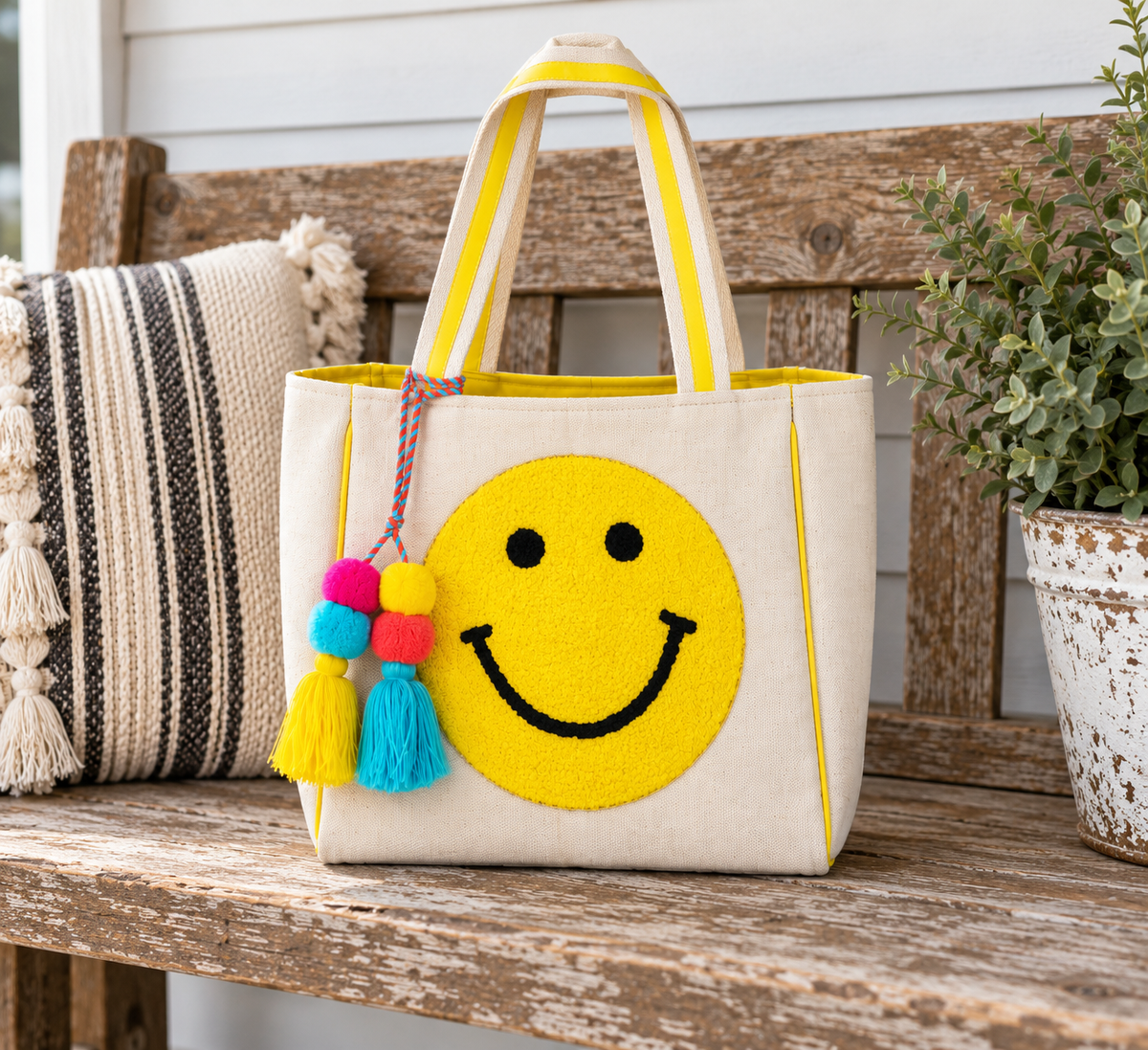 Smiley Face Tote Bag displayed on rustic wooden bench showing natural canvas body with large textured yellow smiley face, yellow striped handles, yellow trim, and colorful pom-pom tassel with striped pillow and potted plant backdrop
