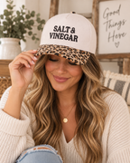 Brunette woman with wavy highlighted hair in cream chunky knit sweater and distressed jeans smiling down while tilting brim of Salt & Vinegar Two-Tone Baseball Cap in Leopard showing cream crown with black embroidered lettering and leopard print bill, in farmhouse interior with eucalyptus in white vase and Good Things Here wood sign in background
