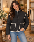 Brunette woman smiling in black quilted contrast trim jacket with cream piping and gunmetal snap buttons, paired with blue jeans in a warm boutique interior with eucalyptus and cream florals
