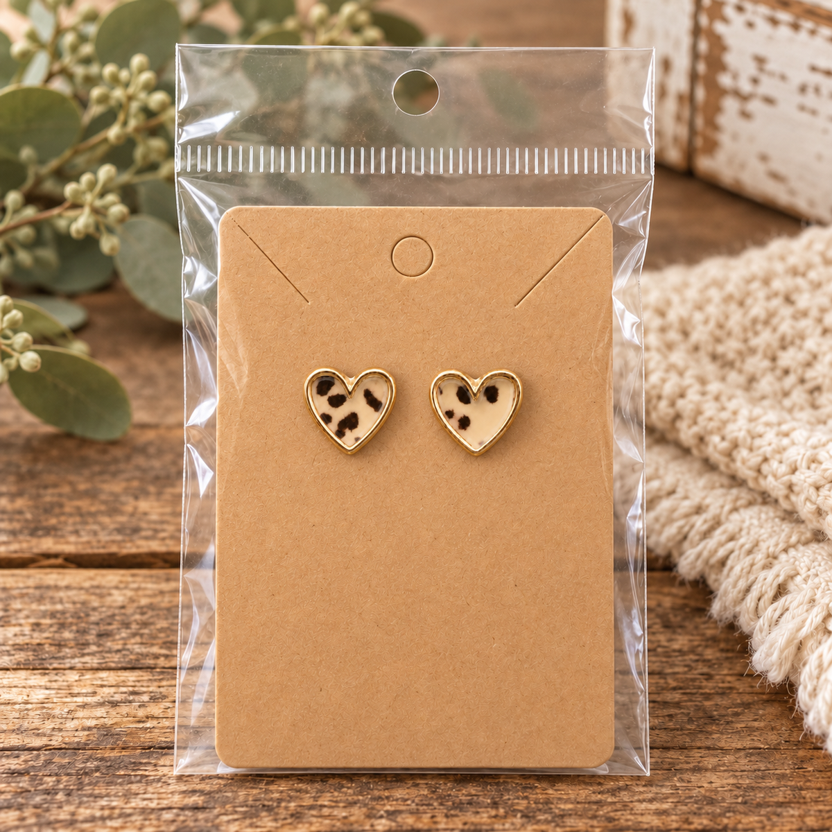 Leopard heart stud earrings with gold-tone trim on kraft card in clear packaging on rustic wood surface with eucalyptus berry sprigs and cream knit
