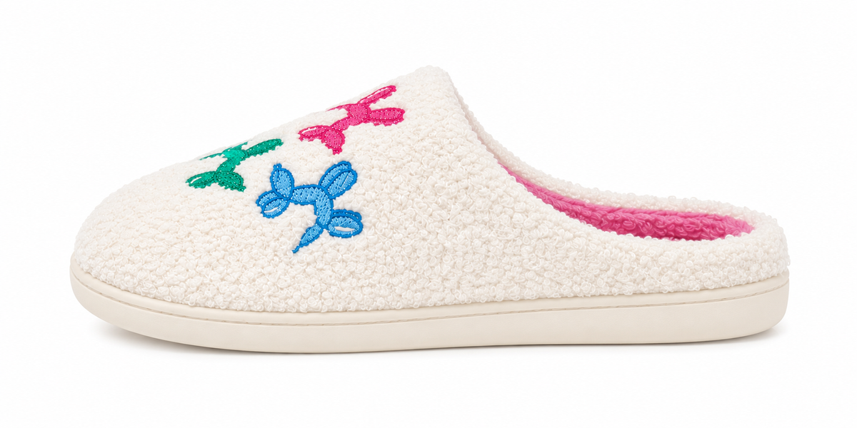 Side profile of one Ivory Balloon Dog Fuzzy Slipper showing ivory fuzzy exterior with colorful embroidered balloon dog accents in pink, blue, and green with bright pink interior lining and cream rubber sole
