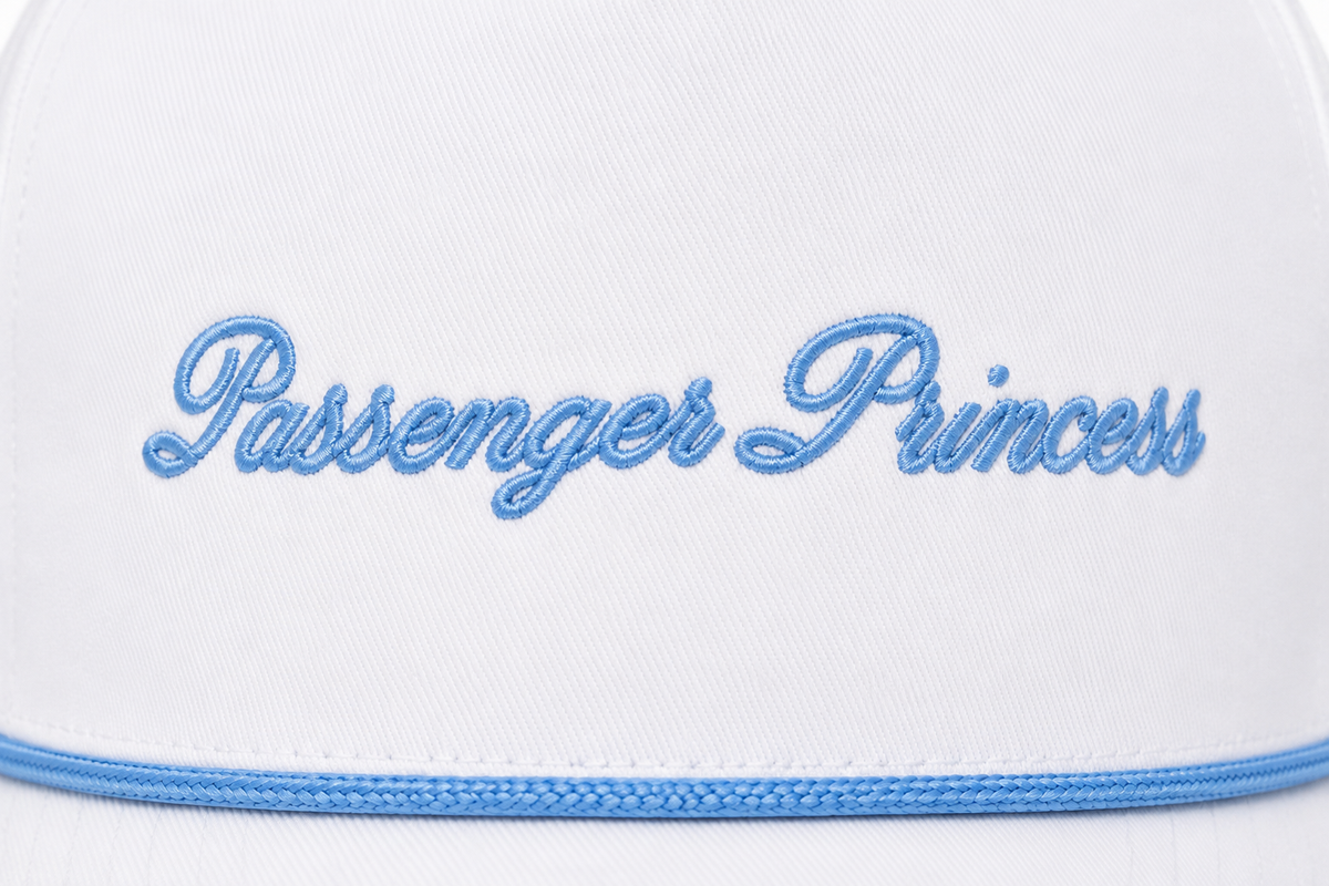 Close-up of light blue embroidered "Passenger Princess" script lettering on a white trucker hat foam front panel with blue rope accent along the brim.
