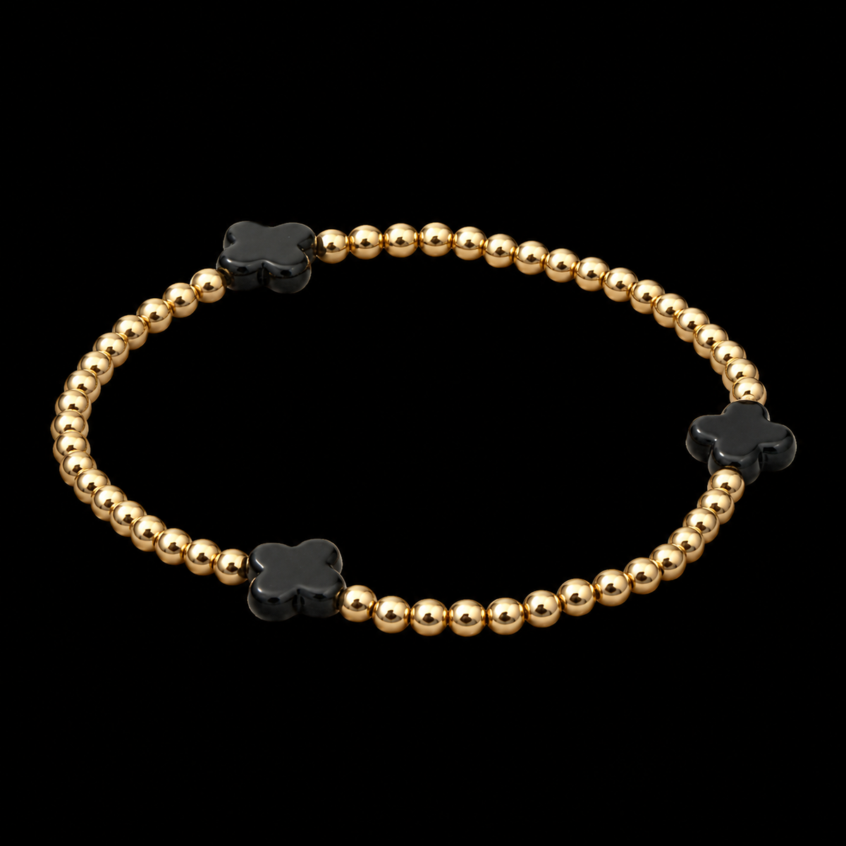 Gold beaded stretch bracelet with black clover accents shown at an angle on black reflective surface
