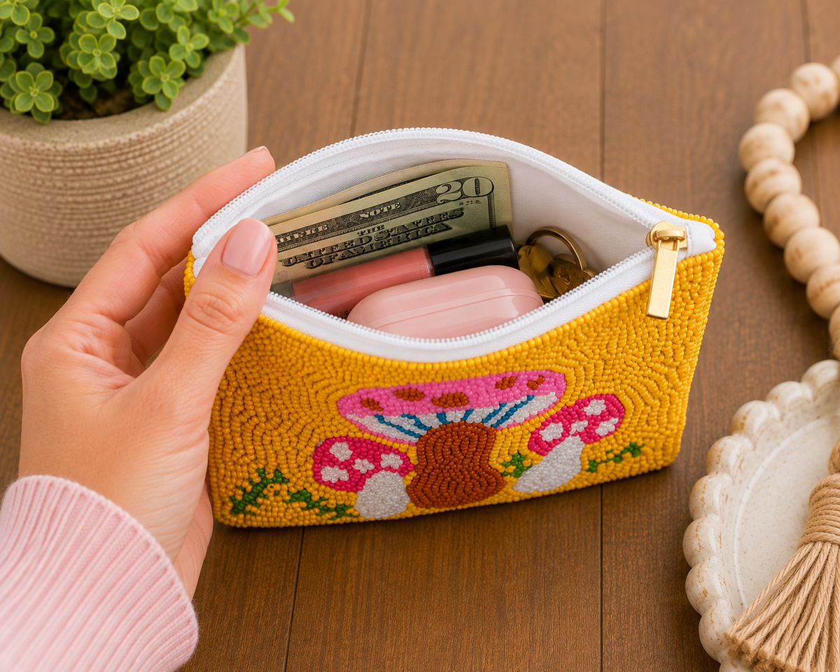 A hand holds an open yellow embroidered zip pouch with a mushroom design, showing it filled with cash, a pink case, and lip balm on a wooden table.