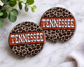 Tennessee Leopard Car Coaster Set — set of 2 ceramic coasters with leopard print and bold orange "Tennessee" block lettering with script overlay, on marble surface
