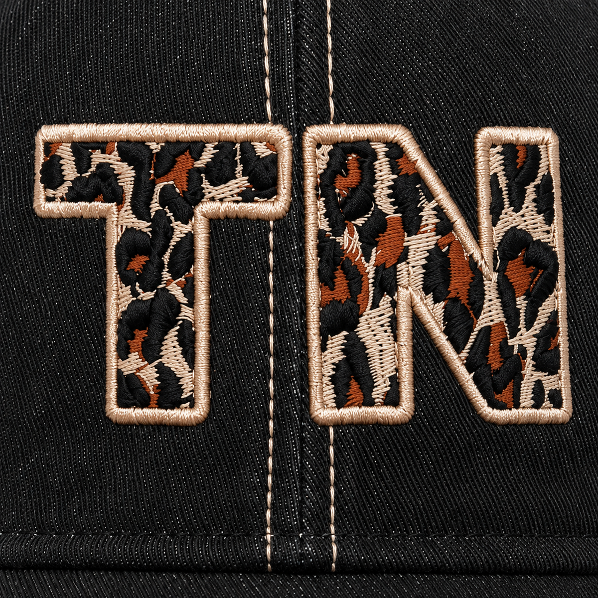 Close-up of bold oversized "TN" embroidery in brown and black leopard print on a distressed black trucker hat front panel.
