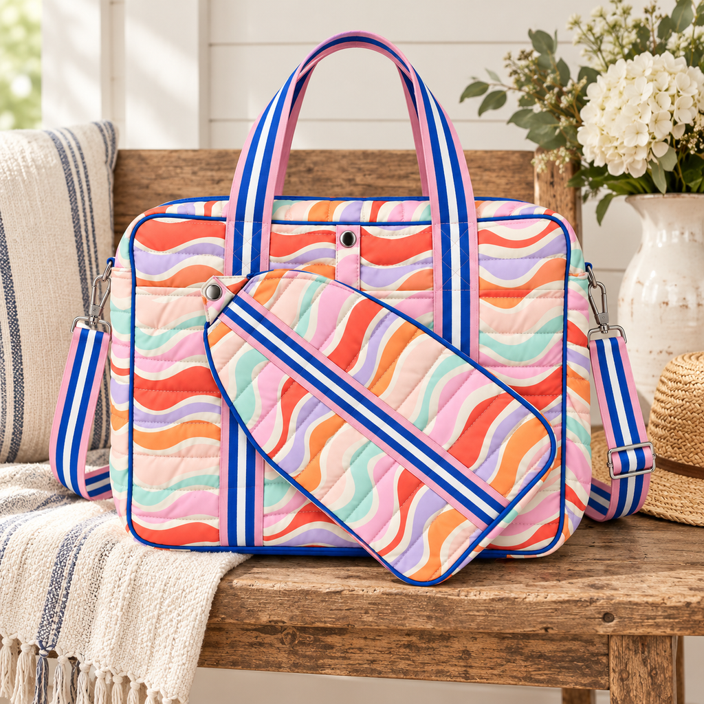 Retro Wave Pickleball Bag and removable paddle holder on rustic wood bench showing multicolor retro wave quilted print with royal blue trim, pink and blue stripe handles, and adjustable blue stripe crossbody strap beside striped gray linen pillow, cream waffle throw, woven straw hat, and white hydrangeas in white vase on white shiplap background
