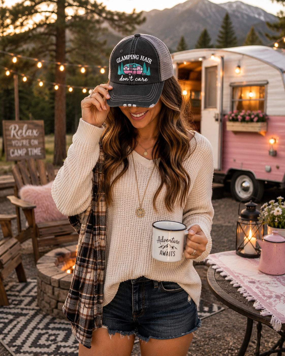 A woman stands outdoors at a campsite wearing a black trucker hat with a colorful camper graphic and the text "Glamping Hair Don't Care," holding a white mug, with a plaid shirt draped over her shoulder.
