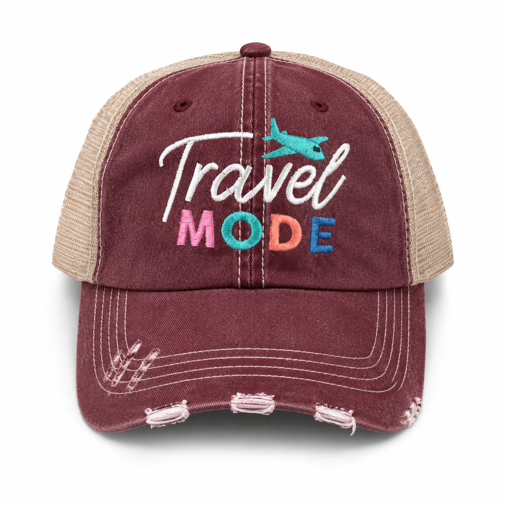 A distressed trucker-style baseball cap in washed burgundy and beige mesh, featuring 'Travel Mode' embroidered on the front with a small teal airplane graphic.