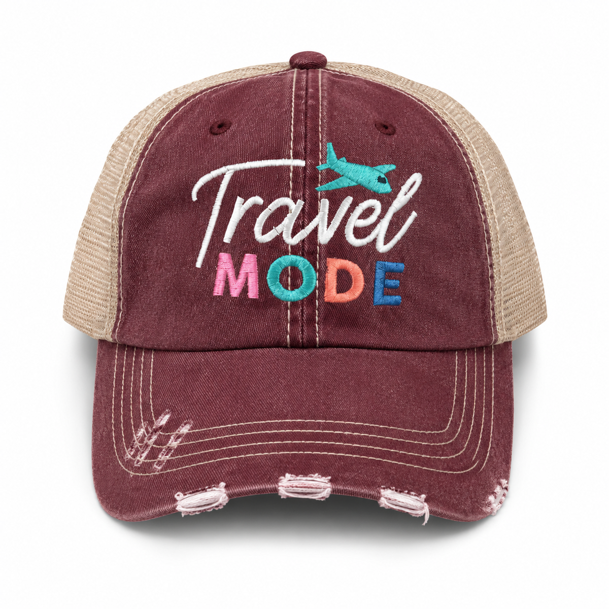 A distressed trucker-style baseball cap in washed burgundy and beige mesh, featuring 'Travel Mode' embroidered on the front with a small teal airplane graphic.